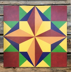 A barn quilt square, made up of different colored and shaped pieces, forming a pleasant pattern